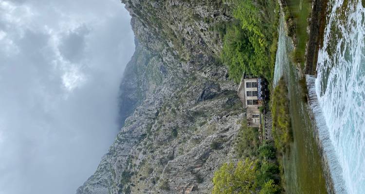 Cañón de montaña con un pequeño edificio de piedra junto a un río caudaloso bajo un cielo nublado