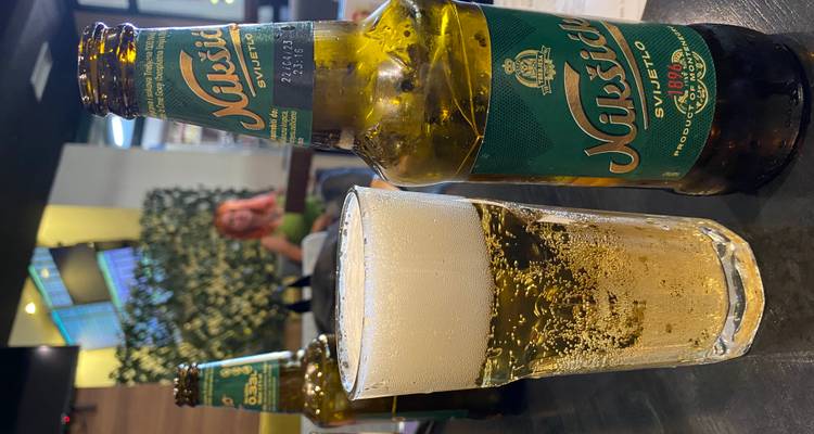 Vaso y botella de cerveza Nikšićko en una mesa de bar con una mujer sentada de fondo