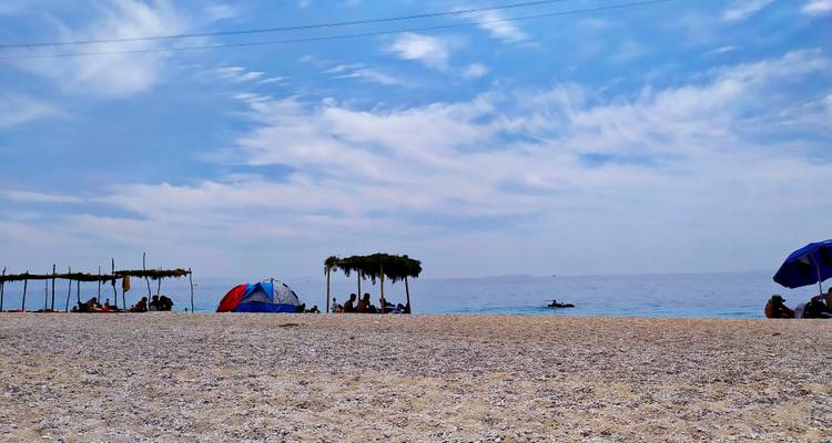 Pebble beach with sunshades, tents and people relaxing by calm blue sea.