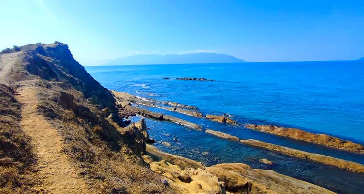 Rocky coastal cliff path beside vivid blue sea with distant hazy mountains.