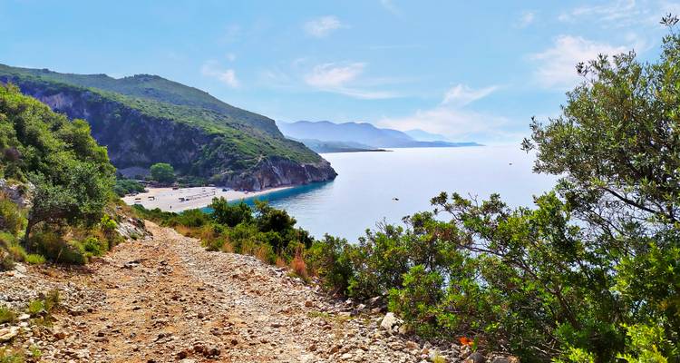 Rocky track descending towards a secluded crescent beach with calm seas and mountains.