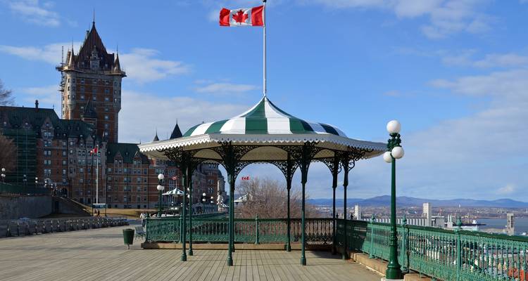 Belvédère victorien sur une promenade en bois surplombant le fleuve avec le Château Frontenac et le drapeau canadien en arrière-plan.