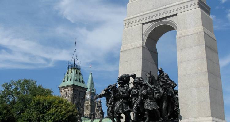 Arc commémoratif de pierre avec des statues de soldats en bronze se dressant devant les tours du parlement gothiques et le ciel bleu d'été.