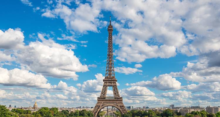 Eiffel Tower dominates a cloud-speckled blue sky rising above green treetops and the Paris skyline.