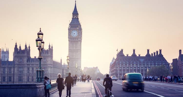 Les piétons et les cyclistes traversent le pont de Westminster à l'aube avec Big Ben en silhouette contre un lever de soleil brumeux.