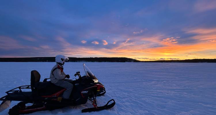 <p>Une personne sur une motoneige sur un vaste lac gelé, silhouettée contre un ciel au coucher de soleil d'un orange éclatant.</p>