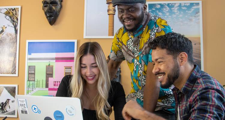 Three young colleagues laughing around a laptop in a room decorated with travel photos.