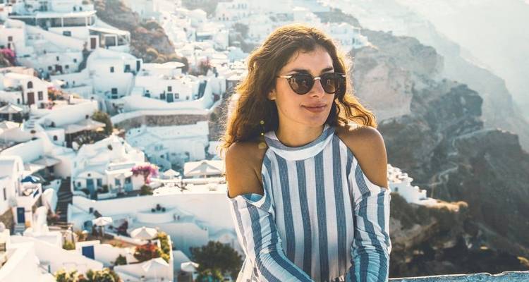 Woman in striped top poses with Santorini’s whitewashed clifftop houses cascading behind her.