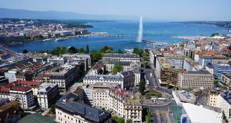 Panoramisch luchtfoto van Genevas waterfront met de Jet d'Eau die uit het Meer van Genève omhoog rijst onder een blauwe hemel.