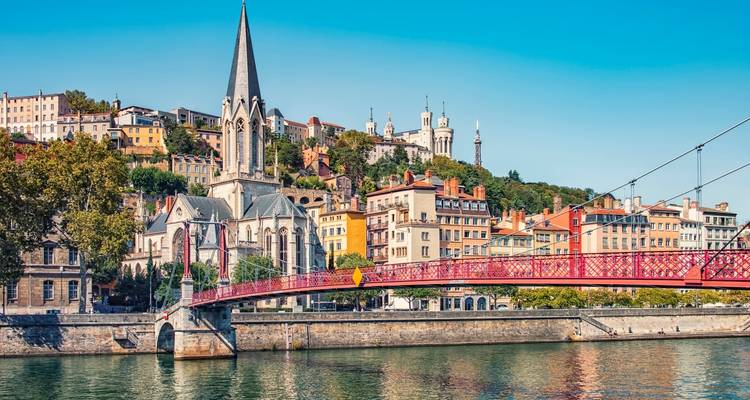 Side view of Lyon’s red Passerelle footbridge with hillside basilica and colourful riverside buildings.