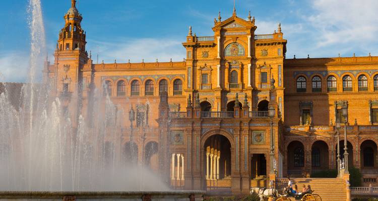 Façade centrale de la Plaza de España avec sa fontaine projetant l'eau haut dans la lumière de l'après-midi et une calèche attendant à proximité.
