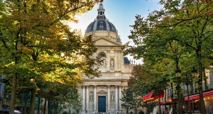 Sun-dappled trees framing the ornate dome of the Sorbonne in a quiet Parisian square.