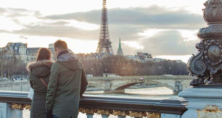 Couple leaning on Pont Alexandre III overlooking the Seine with the Eiffel Tower and dramatic evening clouds.