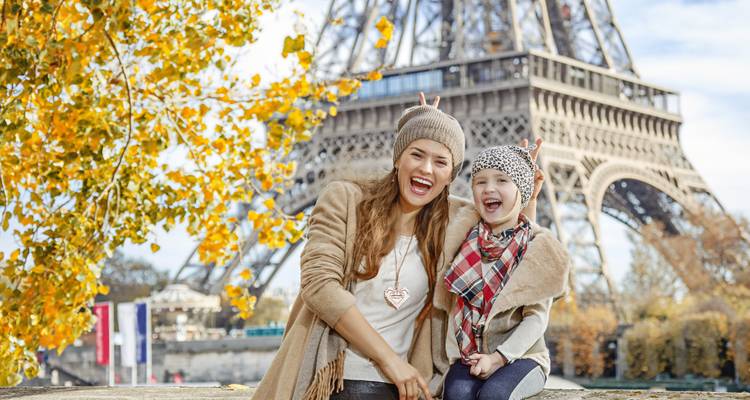 Mother and young daughter laughing in front of the Eiffel Tower amid autumn foliage.