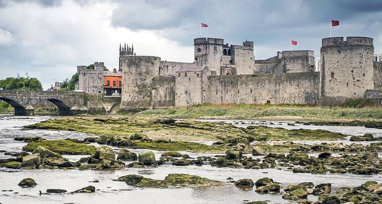 Stone medieval castle with towers and flags beside a rocky riverbed under grey skies