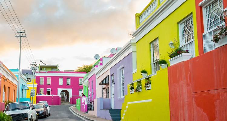 Una calle tranquila bordeada de casas pintadas con colores vibrantes en tonos amarillo, verde, púrpura y rosa bajo un cielo suave.