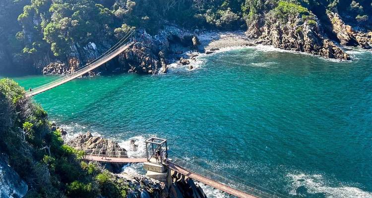 Puente colgante cruzando una bahía de aguas cristalinas azules en la costa de un exuberante parque nacional.