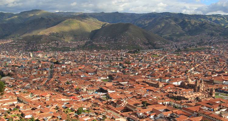 Luchtfoto van het rood-dakige Cusco tussen de groene Andese bergen onder een wolkenlucht.
