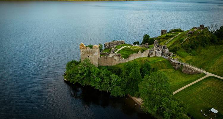 Aerial view of the ruins of Urquhart Castle jutting into the dark waters of Loch Ness with lush greenery around.