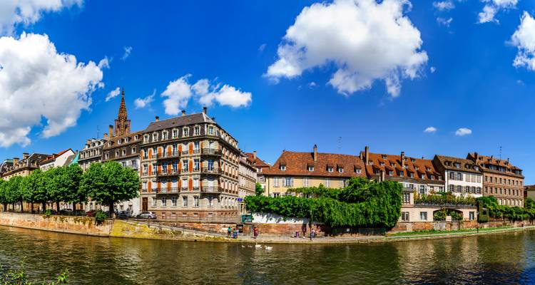 Malerische Flusslandschaft mit historischen Gebäuden und dem hohen Spitzturm der Straßburger Kathedrale, gespiegelt in der Ill unter einem hellblauen Himmel mit vereinzelten Wolken.