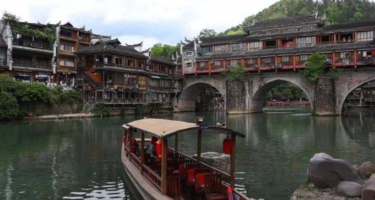 Traditional wooden houses and stone arches span a calm river where a boat with red lanterns carries passengers in Fenghuang.