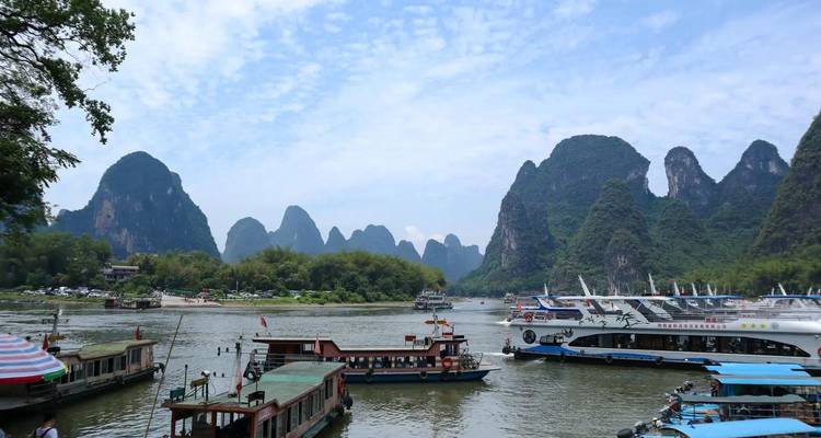 Tour boats wait at a dock on the Li River surrounded by steep karst peaks under a patchy sky.