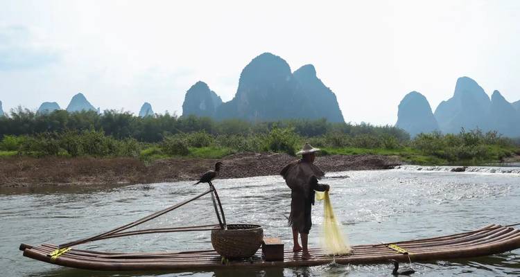 A cormorant fisherman casts a yellow net from a bamboo raft on the Li River framed by silhouetted karst peaks.