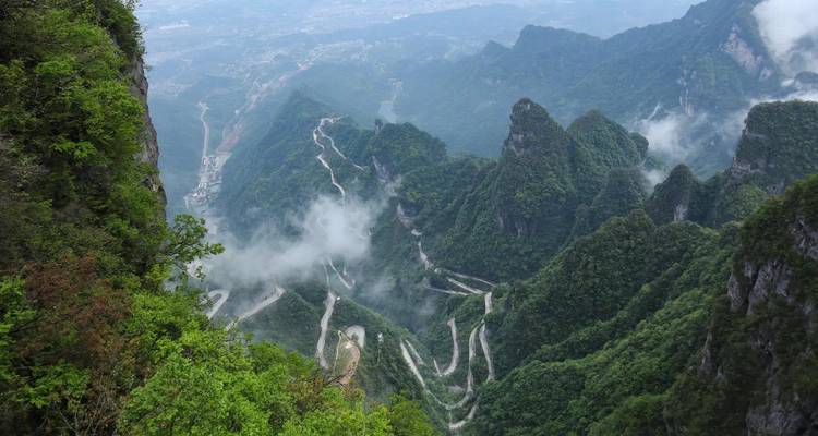 Steep serpentine mountain road climbs through misty green peaks in Zhangjiajie National Forest.