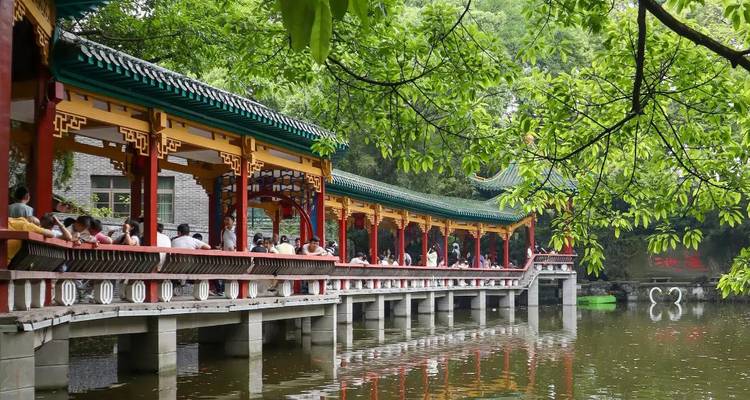 Colourful covered walkway with traditional Chinese architecture stretches over a reflective pond beneath leafy trees.