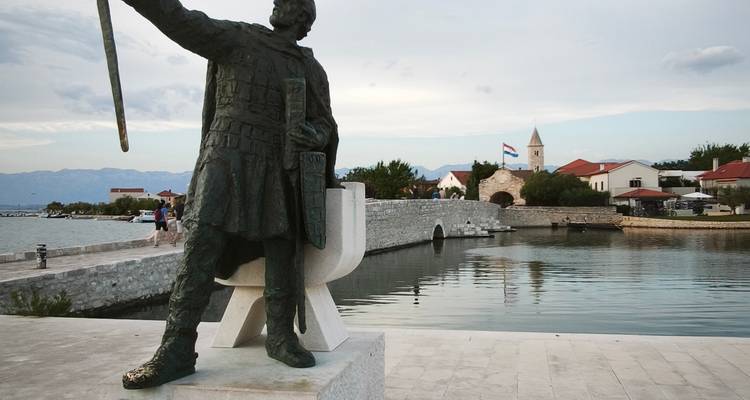 Statue de bronze surplombant un pont de pierre et un port avec le drapeau croate