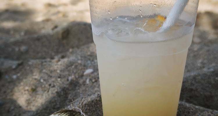 Icy lemonade in a plastic cup with a straw resting on sandy beach ground