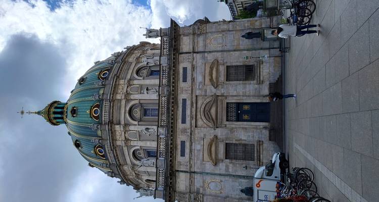 Grande église en marbre avec dôme, statues et toit en cuivre vert, vue depuis une place avec des visiteurs, des bicyclettes et un ciel nuageux au-dessus.