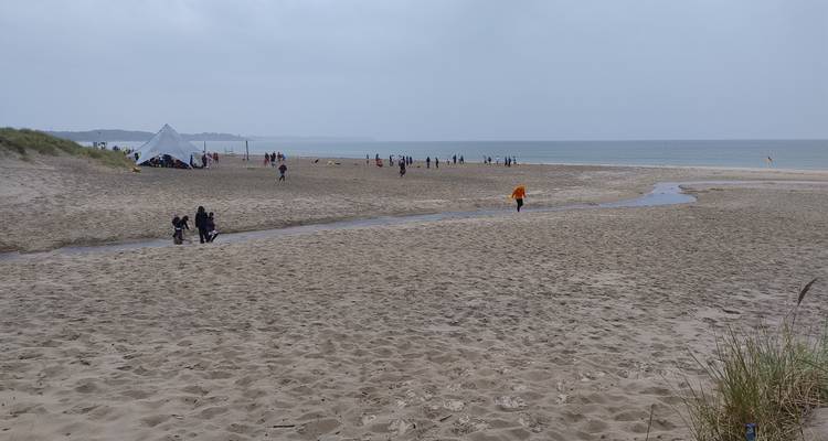 Large plage de sable balayée par le vent avec des visiteurs dispersés et un ruisseau peu profond rencontrant la mer du Nord grise par une journée nuageuse.