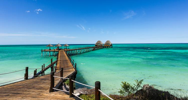 A wooden pier stretches over clear turquoise sea toward thatched huts under a bright blue sky.