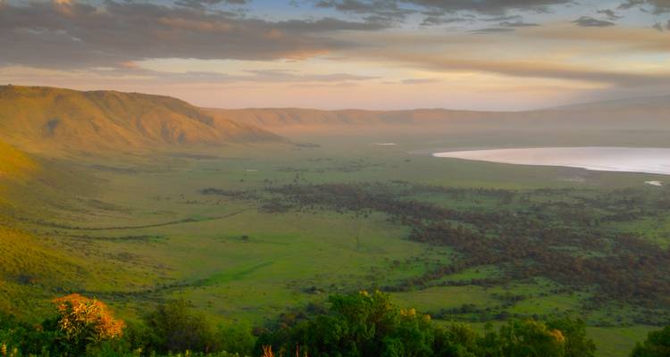 A panoramic view into a lush green crater with a distant alkaline lake and surrounding cliffs lit by soft morning light.