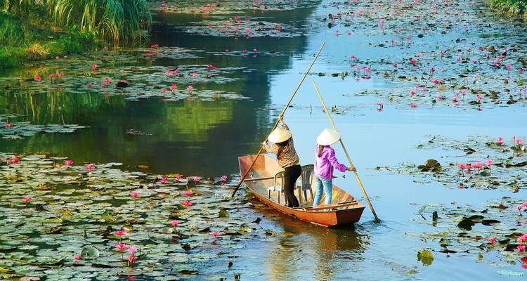 Deux habitants font avancer une barque en bois à l'aide de perches à travers un étang recouvert de nénuphars roses.