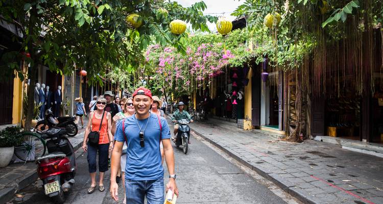 Les touristes se promènent dans une rue décorée de lanternes, bordée de bougainvillées et de maisons-boutiques historiques, tandis que des cyclomoteurs les dépassent.