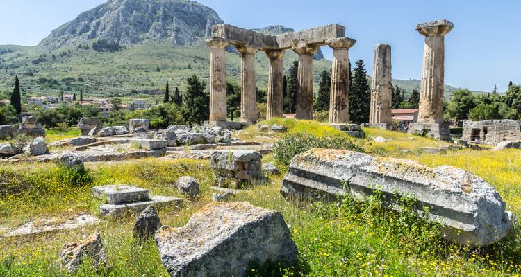 Ruins of the ancient Temple of Apollo in Corinth with wildflowers and a rocky mountain backdrop.