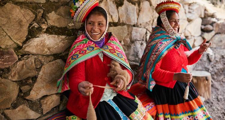 Deux femmes andines vêtues de costumes traditionnels colorés filant la laine près d'un mur en pierre.