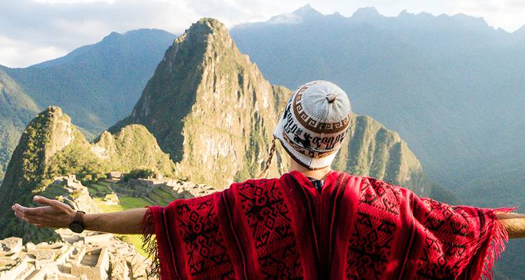 Voyageur en poncho rouge et chapeau andin les bras écartés surplombant le Machu Picchu.