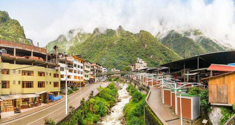 The riverside town of Aguas Calientes nestles beneath misty green Andean peaks with a rushing stream at its center.