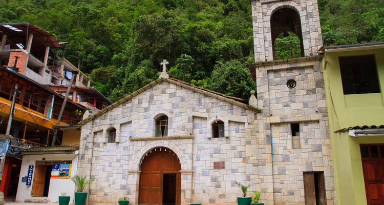 A small stone church with a wooden door and bell tower stands against a lush jungle hillside.
