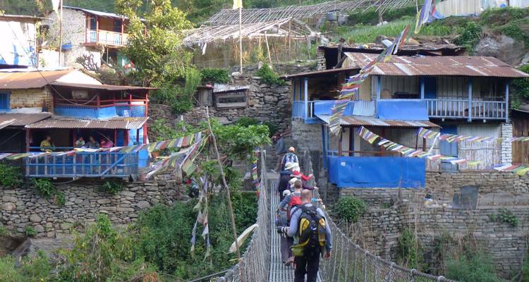Les trekkers traversent un pont suspendu étroit orné de drapeaux de prière entre des maisons rustiques.