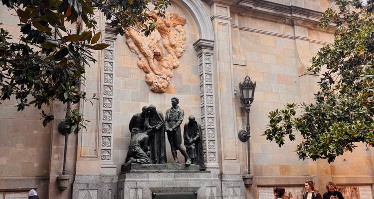 Mur en pierre avec statue historique et visiteurs conversant sous des branches feuillues dans le Quartier Gothique de Barcelone.