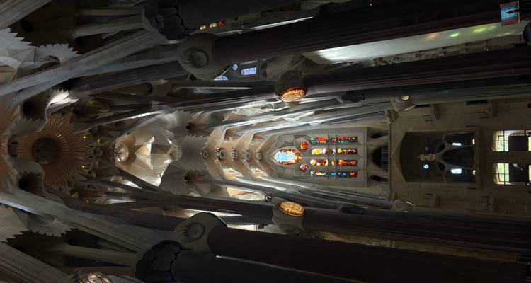 Nef centrale faiblement éclairée de la Sagrada Familia avec des colonnes élancées et une fenêtre en vitrail focal imposante.
