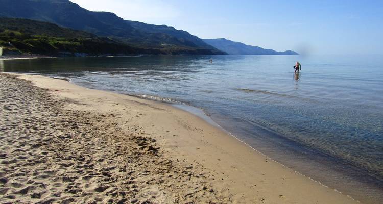 Ruhiger Sandstrand mit sanften Wellen, an dem ein einsamer Badegast im flachen Wasser steht.