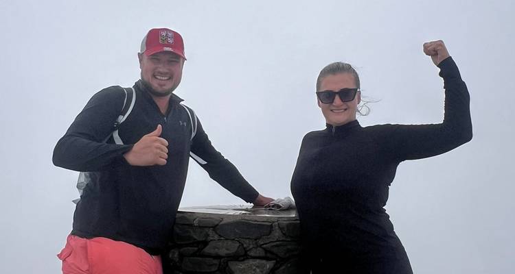 Two hikers posing with thumbs-up at the misty summit trig point of Mount Snowdon.