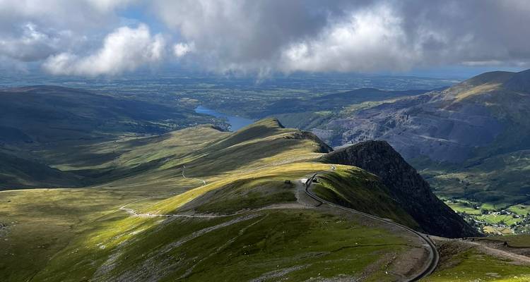 Sweeping aerial ridge view over Snowdonia with sunlit valleys, winding path and distant lake.