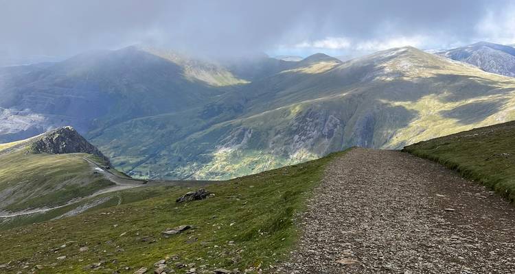 Gravel ridge path descending through dramatic Snowdonia mountain scenery under patchy clouds.