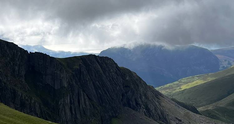 Rugged dark cliff face under moody clouds with distant misty peaks in Snowdonia.